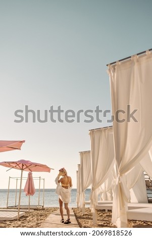 Slim caucasian girl standing back to camera in distance, facing sea on beach. Chic blonde with short hair in swimsuit, shirt top and black sandals. Concept of solitude with nature.
