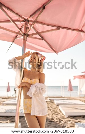 Gentle image of pretty young woman standing on beach holding pink beach umbrella. Blonde woman with colored bandage on her head is wearing yellow swimsuit and white shirt.