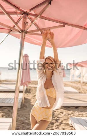 Happy young woman smiling with teeth stretching her arms above her head under beach umbrella. Blonde in yellow swimsuit and white shirt stands on seashore. Concept of solitude with nature.