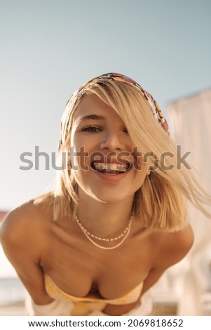 Close-up of caucasian young sweet lady with snow-white even smile against background of sky. Blonde with bob haircut in swimsuit with pearl necklace and kerchief on her head.