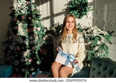 A beautiful young woman with blond hair in light clothes sits on the sofa and holds a wrapped gift with a bow in her hands. Time to open Christmas presents. New Years is soon