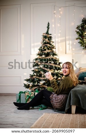 Pretty young woman with blond hair sits near a Christmas tree with coffee. New Year is coming soon. Christmas atmosphere in a cozy home