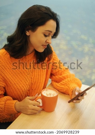 Smiling young woman in orange sweater with croissant and cup of coffee posing on cafe background