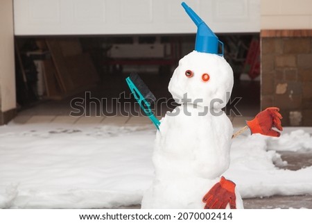 Snowman motorist stands in front of the garage. A watering can, a brush and work gloves are used for decoration

