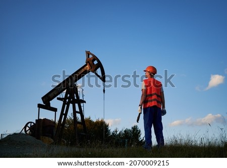 Back view of male worker in protective helmet standing near petroleum pump jack under blue sky. Petroleum engineer in work vest holding industrial wrench and looking at oil pump rocker-machine.
