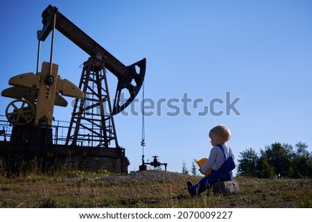 Rear view of preschool guy sitting on one stone, holding construction helmet in hands. Male kid looking at operation of drilling rig station for oil extraction outdoors under blue sky.