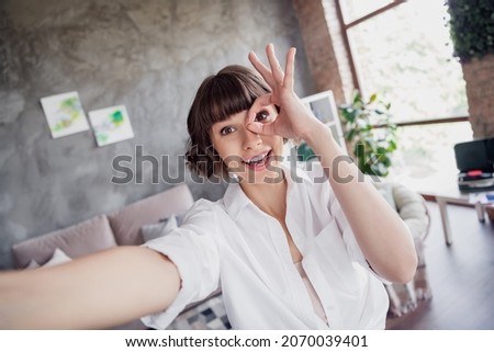 Photo of cheerful positive amazed woman make finger okay sign eye look make selfie indoors inside house home