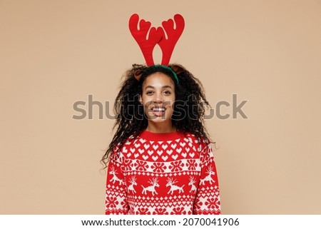 Smiling young african Santa woman in Christmas sweater fun decorative deer horns on head isolated on pastel beige background studio portrait. Happy New Year celebration merry x-mas holiday concept