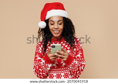 Fun young african american Santa woman in Christmas hat sweater hold in hand use mobile cell phone isolated on pastel beige background studio portrait. Happy New Year 2022 merry x-mas holiday concept