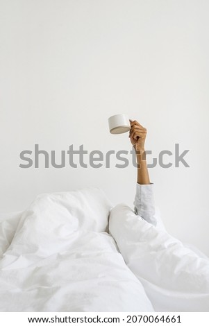 Concept of early morning wake up. Vertical view dark skinned woman in pajamas on bed, wrapped in warmth cozy blanket, raise hand with coffee cup. Female girl holding mug against white copy space wall Royalty-Free Stock Photo #2070064661
