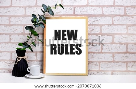 On a white table against a light brick wall, a branch of eucalyptus in a dark vase, a white cup and a light wooden frame with the text NEW RULES. Home office interior.