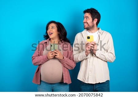 young couple expecting a baby standing against blue background holding a smartphone and looking sideways at blank copyspace.