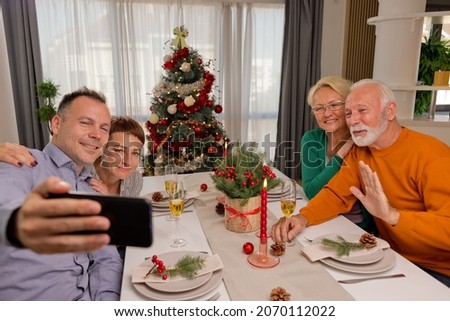 Happy family or friends sitting by nicely decorated Christmas table and taking picture together or having video call using smart phone during festive season