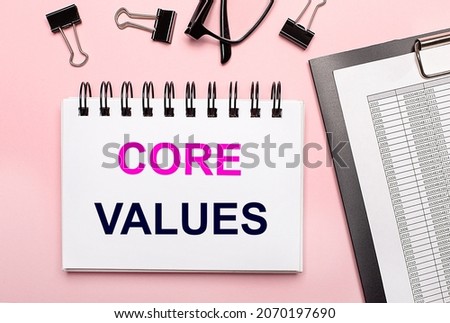 On a pink background, reports, black paper clips, glasses and a white notebook with the text CORE VALUES