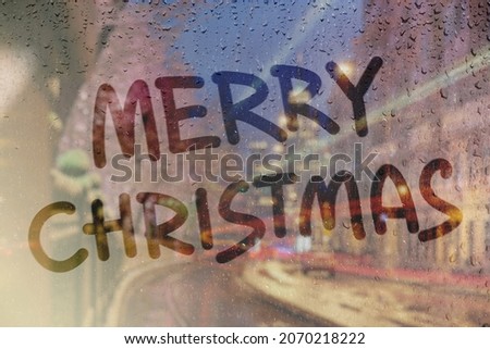 Rainy weather, hand drawn inscription Merry Christmas on the sweaty glass. Night city street with lights on blurred background