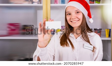 Beautiful young girl office manager in the company smiling wearing the Santa cap having the badge on her white shirt and holding in her hand holiday voucher, copy space.