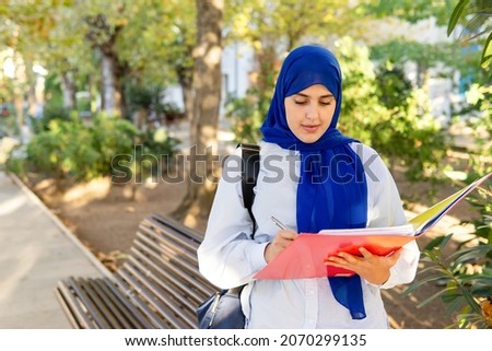 Smiling young Muslim woman in hijab holding a folder and writing in the university campus