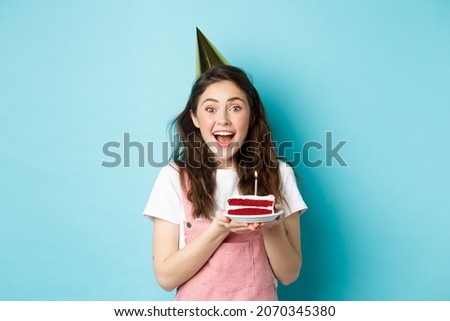 Holidays and celebration. Cheerful birthday girl in party hat holding bday cake and smiling, making wish on lit candle, standing against blue background