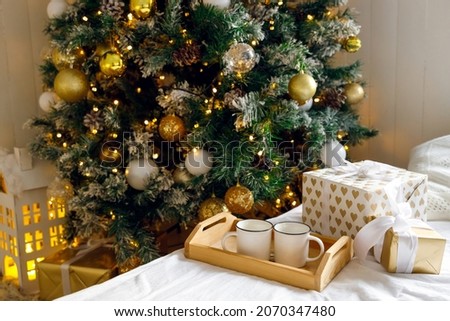 Gift box, wooden tray and two white mugs on the bed on a white sheet, near a decorated Christmas tree. Cozy morning. Coffee in bed