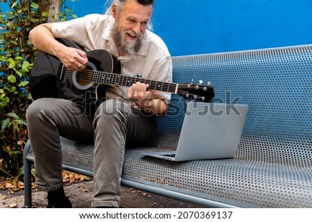 Mature caucasian man sitting on a bench learning to play the guitar with online classes using laptop.