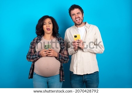 Photo of astonished crazy young couple expecting a baby standing against blue background hold smartphone dislike feedback concept