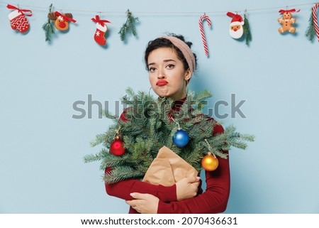 Dissatisfied young brunette woman 20s wears red sweater hold bouquet of spruce branches isolated on plain pastel light blue background studio portrait. Happy New Year 2022 celebration holiday concept