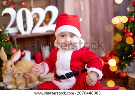 A small child is sitting on a sled under the Christmas trees. A large portrait. A baby in a red Santa Claus costume with a gift in a gift box. Happy Holidays, New Year. Christmas Time