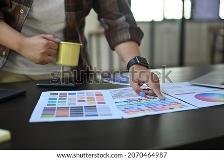Cropped image of a designer hand holding a coffee cup and pointing to the color swatch on the working desk.