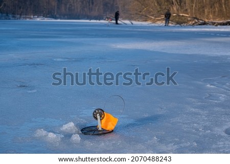 tip-up fishing rod with orange signal flag in sunlight, ready for pike bite in a frozen river hole, fisherman figure, popular seasonal outdoor hobby