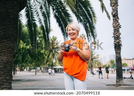 Adult chubby female tourist in stylish casual clothes standing in street near tropical plants with photo camera while looking at camera