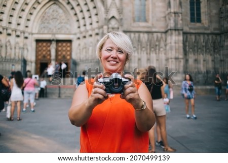 Cheerful plump female tourist in casual clothes standing near old cathedral with photo camera in hands and looking at camera