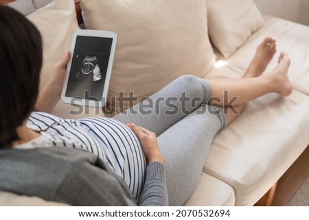 Asian pregnant woman lying on sofa looking at ultrasound scan photo on digital tablet. Concept of pregnancy, Maternity prenatal care.