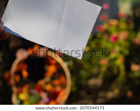 A sheet of white blank paper on a background of multi colored nature. Use it to place text on an empty space. In The Hands Of A Clean White Cardboard On The Background Of A Blurry Colorful Garden.