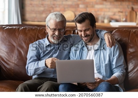 Family time with grown son. Elder and younger men relatives sit on sofa at home hug look on laptop screen discuss online news. Retired dad interested in pc talk ask adult child question about app work