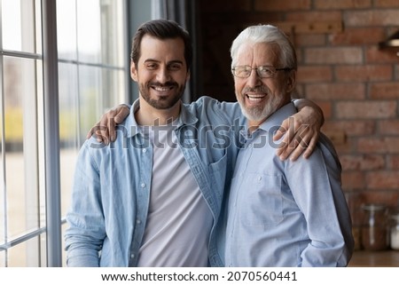 Warm relationship. Family portrait of mature father young adult son stand by window hug shoulders look at camera. Friendly aged grandfather grown grandkid feel emotional bonding enjoy pastime together Royalty-Free Stock Photo #2070560441