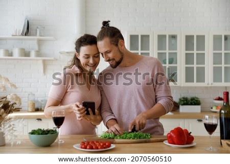 Happy young beautiful woman holding cellphone in hands, showing video with food recipe or sharing funny photo content with laughing husband, enjoying cooking together in modern kitchen on weekend.