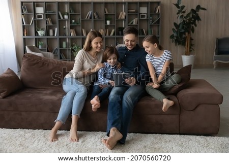 Happy family with children using tablet together, relaxing on cozy couch at home, smiling parents with daughter and son spending leisure time holding device, having fun online, looking at screen