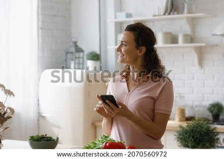 Distracted from cooking healthy food smiling young dreamy woman using cellphone, feeling inspired searching recipe online, preparing for romantic home dating or domestic party in modern kitchen.
