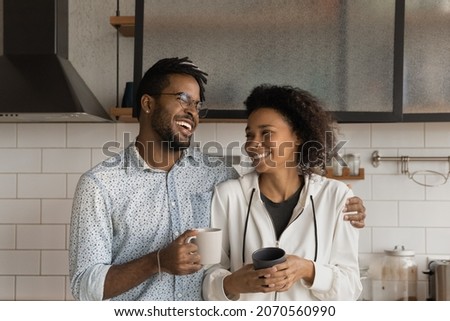 Laughing affectionate young African American family couple drinking morning coffee, standing in modern kitchen, talking speaking joking on weekend at home, spending leisure carefree time together. Royalty-Free Stock Photo #2070560990