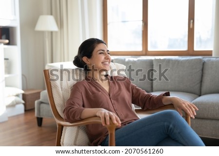 Happy Indian ethnicity woman relaxing on comfy wooden armchair spend time in cozy living room, smile enjoy carefree weekend and comfort furniture, breath fresh conditioned air inside modern apartment Royalty-Free Stock Photo #2070561260