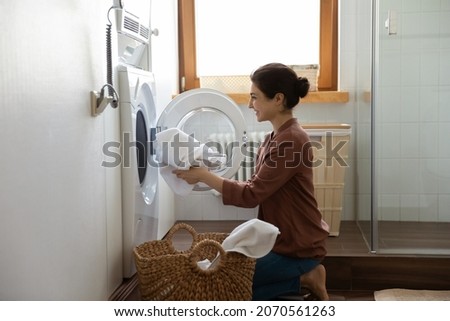 Young happy Indian housewife doing housework putting clothes into washing machine in cozy domestic laundry room. Routine house work, housekeeping, washer-dryer modern appliance or detergent ad concept Royalty-Free Stock Photo #2070561263