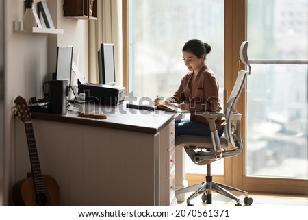 Serious young Indian female sit on ergonomic comfy chair at desk holds pen jotting information, student studying, business woman makes useful notes on paper notebook. Modern office, workflow concept Royalty-Free Stock Photo #2070561371