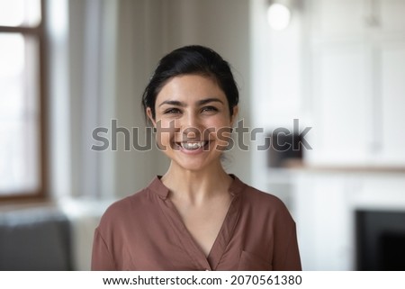 Head shot attractive young 30s Indian female with wide toothy smile posing on camera while standing alone in modern cozy living room at own or rented apartment. Tenancy, housekeeper portrait concept Royalty-Free Stock Photo #2070561380