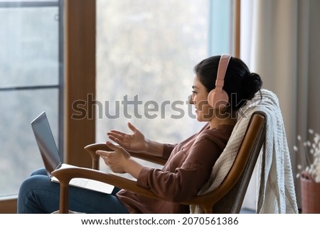 Side view Indian woman sit on armchair wear headphones use laptop talk via video conference enjoy pleasant conversation to family or friend, provide counsel support remotely, video call event concept Royalty-Free Stock Photo #2070561386
