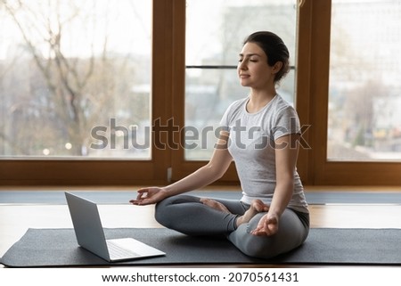 Serene Indian ethnicity woman sit cross-legged on yoga mat listen soothing music on laptop, do meditation practise with eyes closed alone indoor. Remote e coach training, modern tech, work out concept Royalty-Free Stock Photo #2070561431