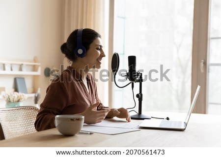 Young Indian ethnicity woman sit at table wear wireless headphones recording podcast for internet audience on laptop makes speech using microscope. Streaming services, on-line radio station concept