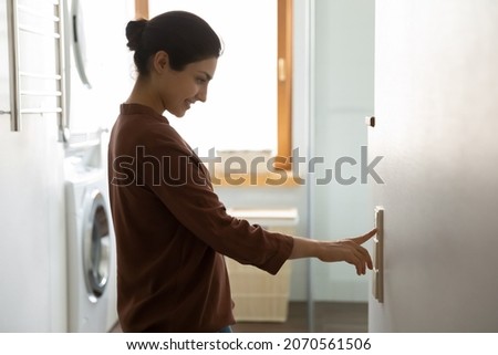 Young Indian ethnicity female push button turns off on light inside modern domestic laundry room, adjusting thermostat for controlling air conditioner and heater inside house. Climate control concept Royalty-Free Stock Photo #2070561506