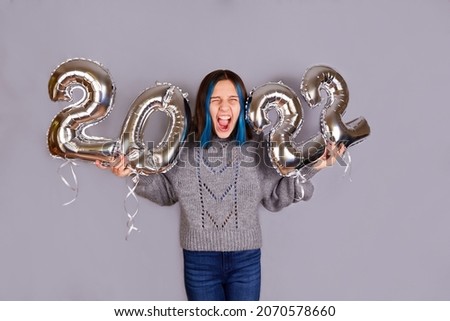 A beautiful emotional girl on a gray background holds silver balloons from the number 2022 in her hands. The brunette closed her eyes with delight from the upcoming holiday. Happy New Year 2022.