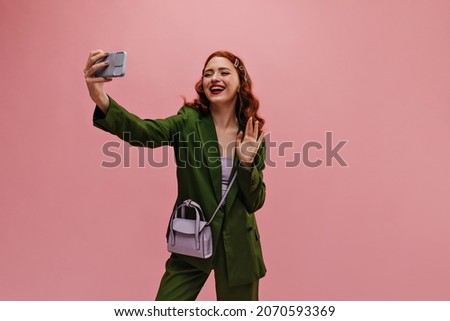 Lovely young woman affably waves into phone screen while chatting on video communication. Caucasian red-haired beauty in business green suit with bag over her shoulder on pink background.