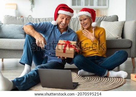 Distance Christmas. Cheerful Senior Couple Making Video Call Using Laptop, Celebrating Holiday Online Wearing Santa Hats And Holding Xmas Gift At Home. Winter Holidays Season, Remote Celebration